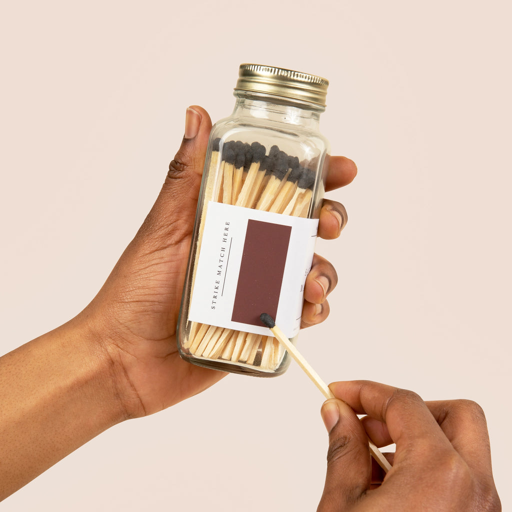 Hand holding a glass match jar with matches and a lit match against a beige background