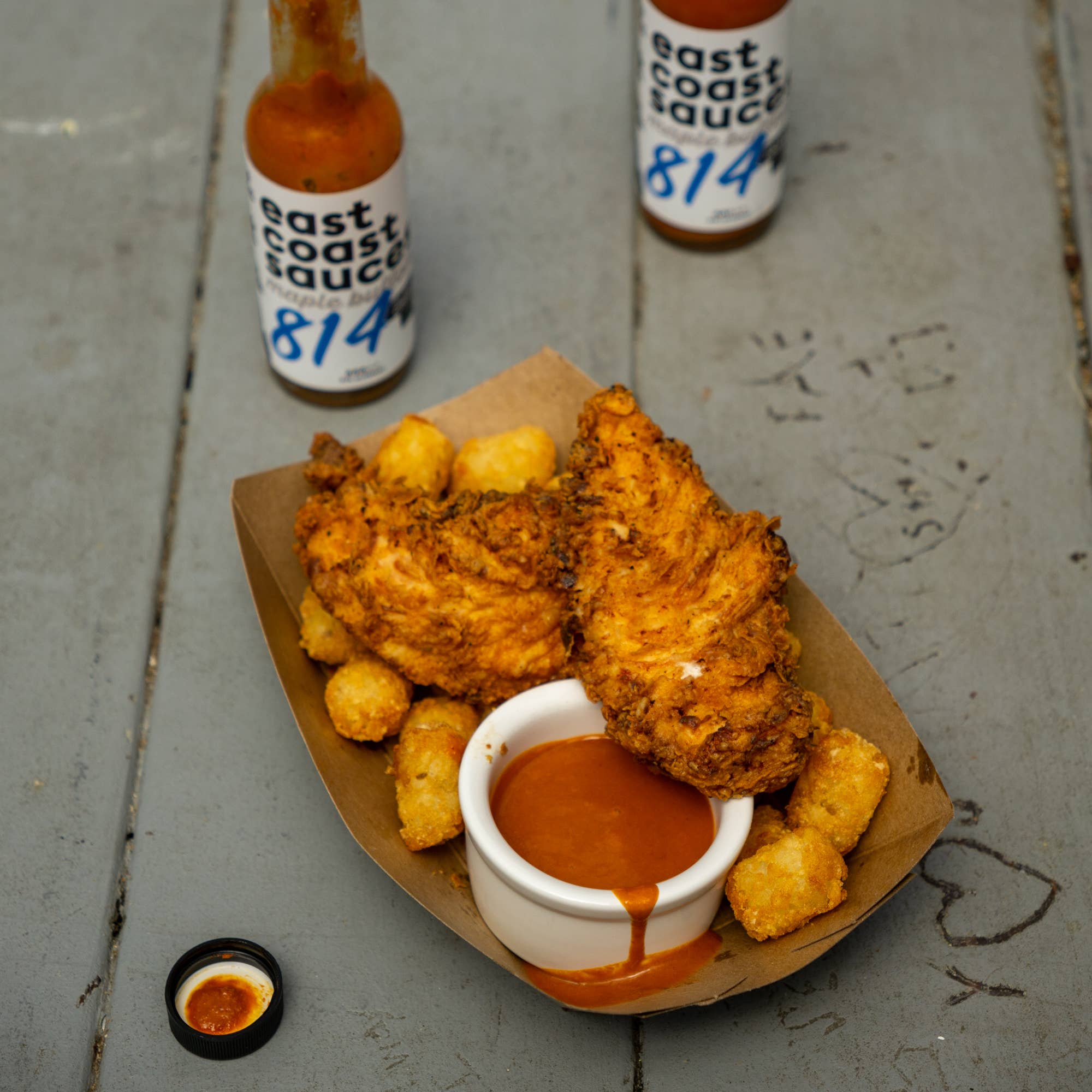Fried chicken and tater tots with a dipping sauce on a paper tray, accompanied by 'east coast sauce' bottles.