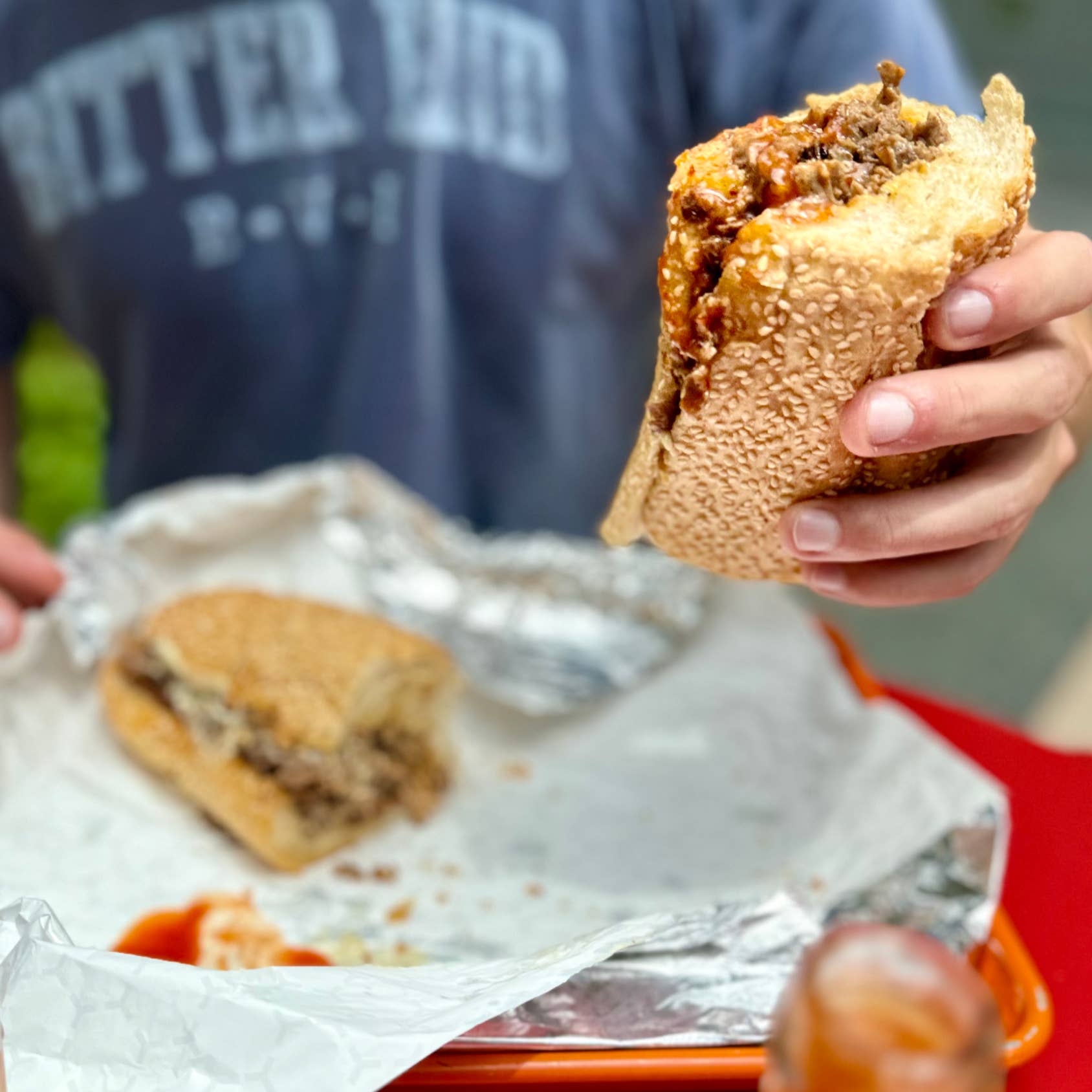 Person holding a deep-fried sandwich with a bite taken out of it, with another sandwich on a tray in the background.