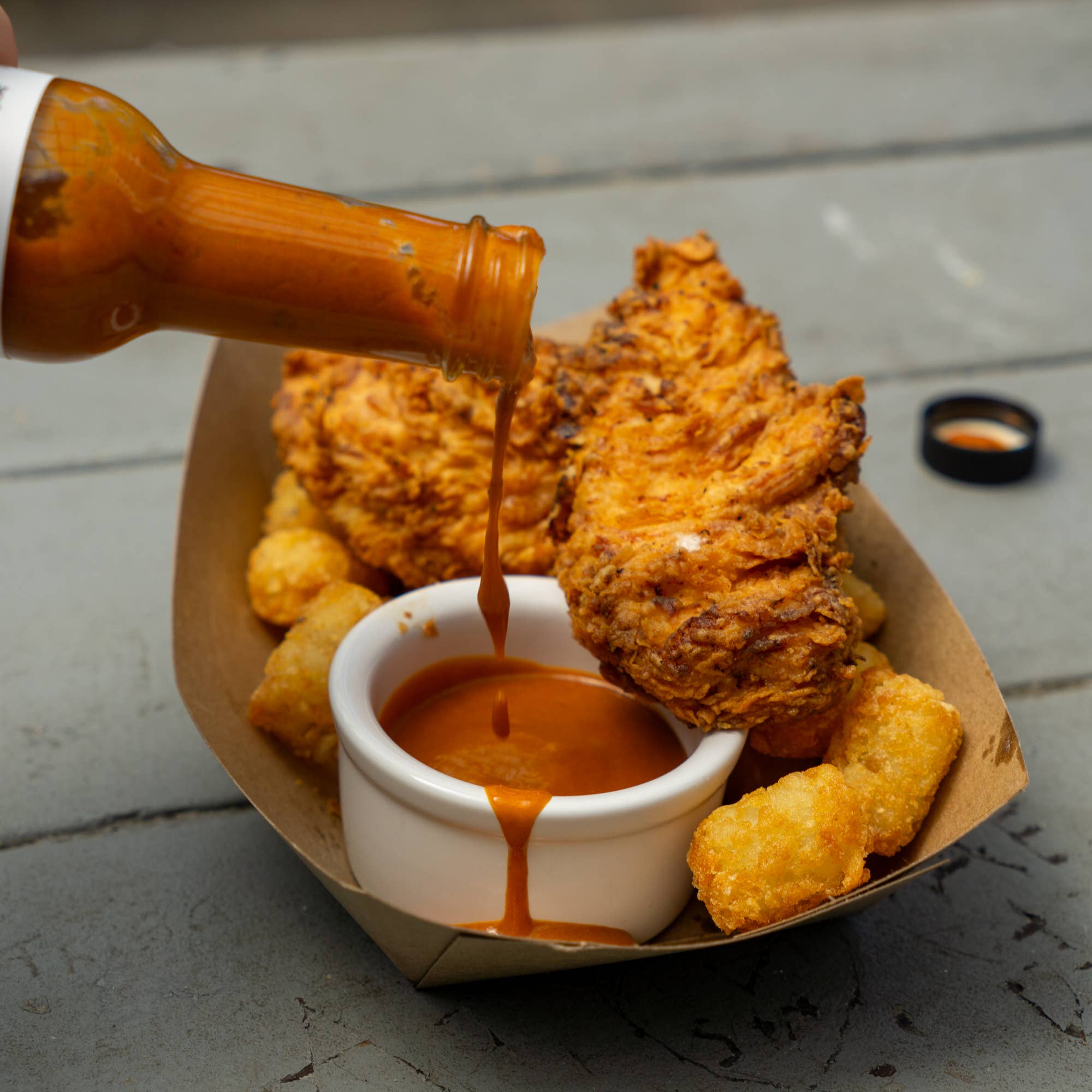 Fried chicken and tater tots with a bottle pouring sauce into a small bowl.