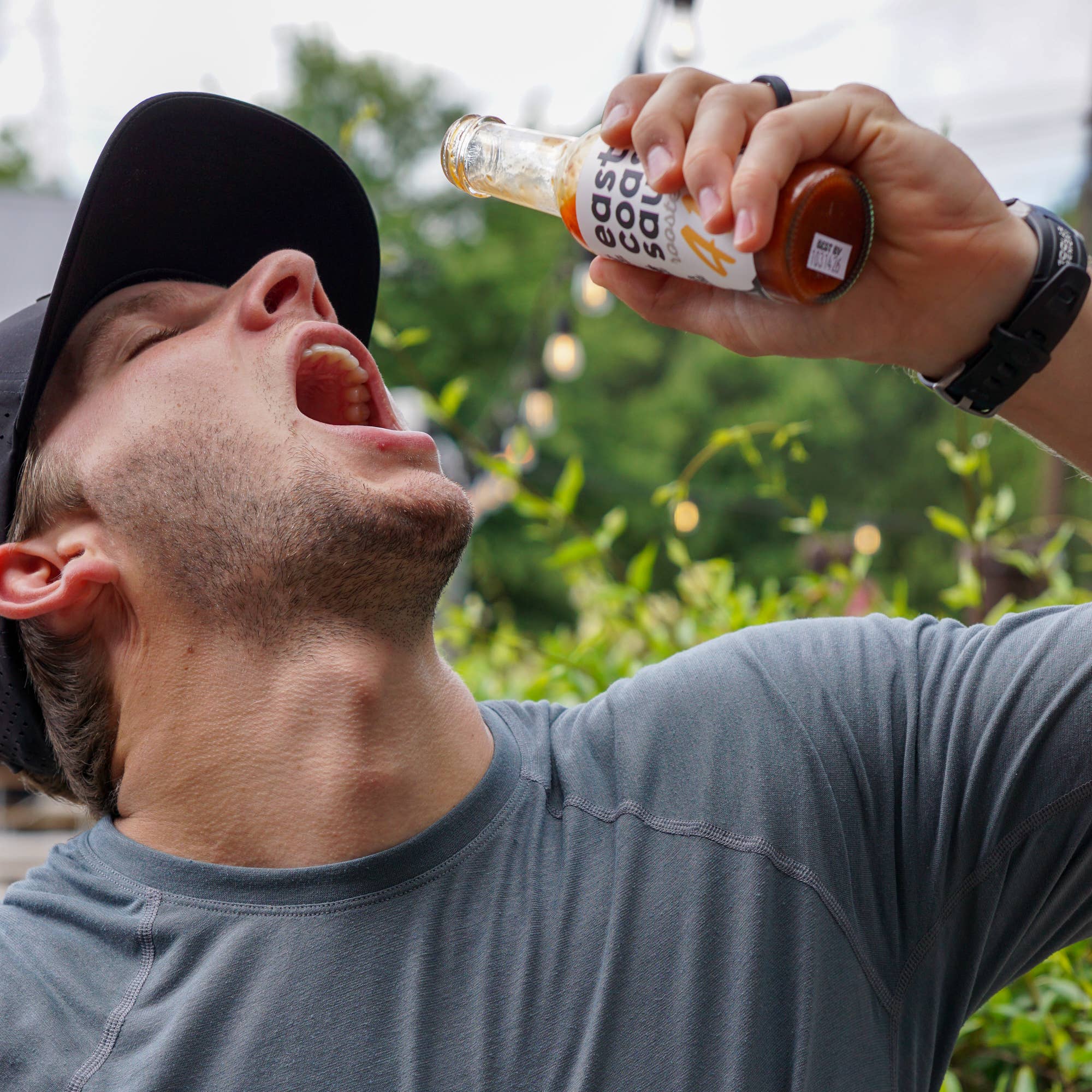 Person drinking from a bottle labeled 'East Coast Sauces' with a blurred outdoor background