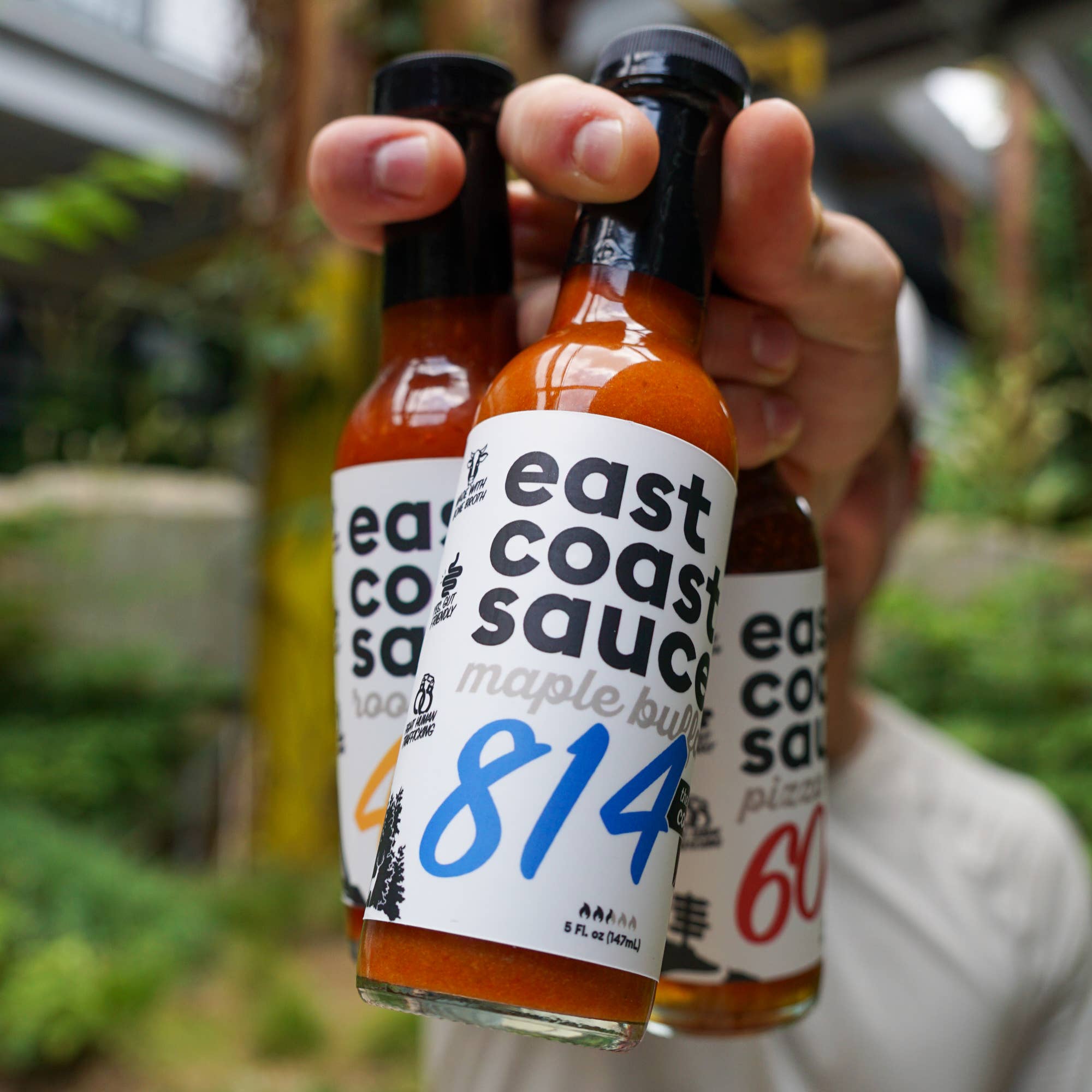 Three bottles of East Coast Sauce held by a person with a blurred outdoor background