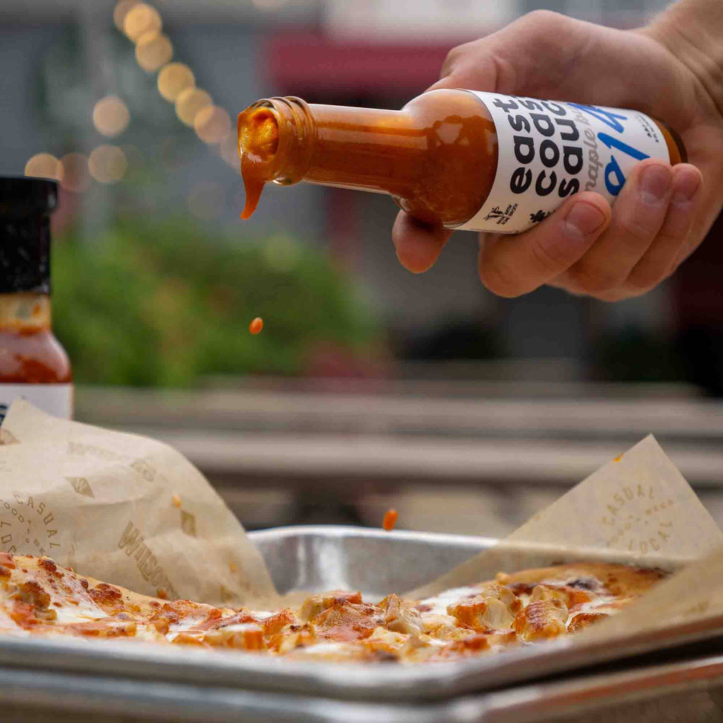 Person pouring hot sauce onto a pizza with a blurred outdoor background