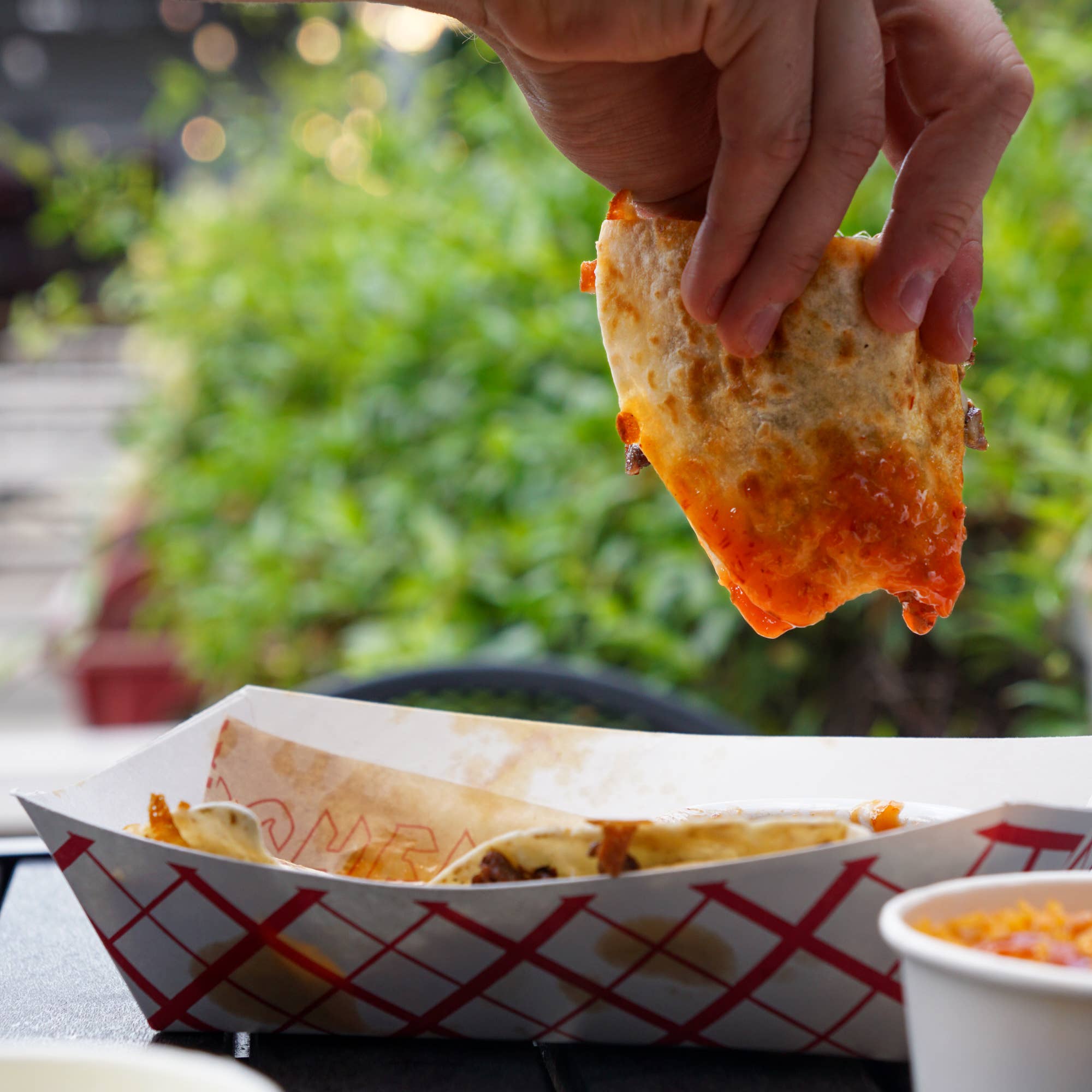 Hand holding a cheesy breadstick over a paper container with more breadsticks and a side dish outdoors.
