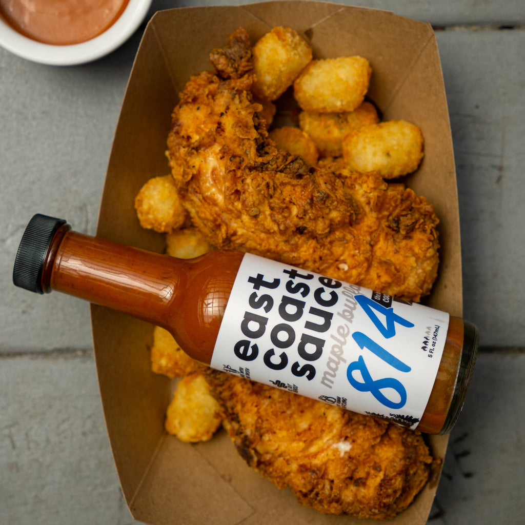 Fried chicken and tater tots in a cardboard container with a bottle of 'east coast sauce by 814' on top.