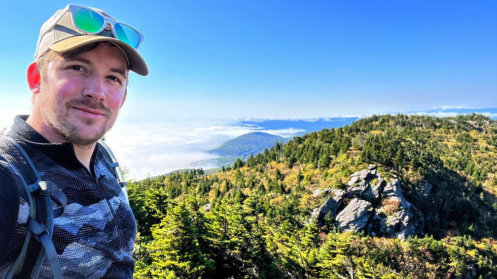 Man taking a selfie on a mountain with a scenic view of trees and mountains in the background.