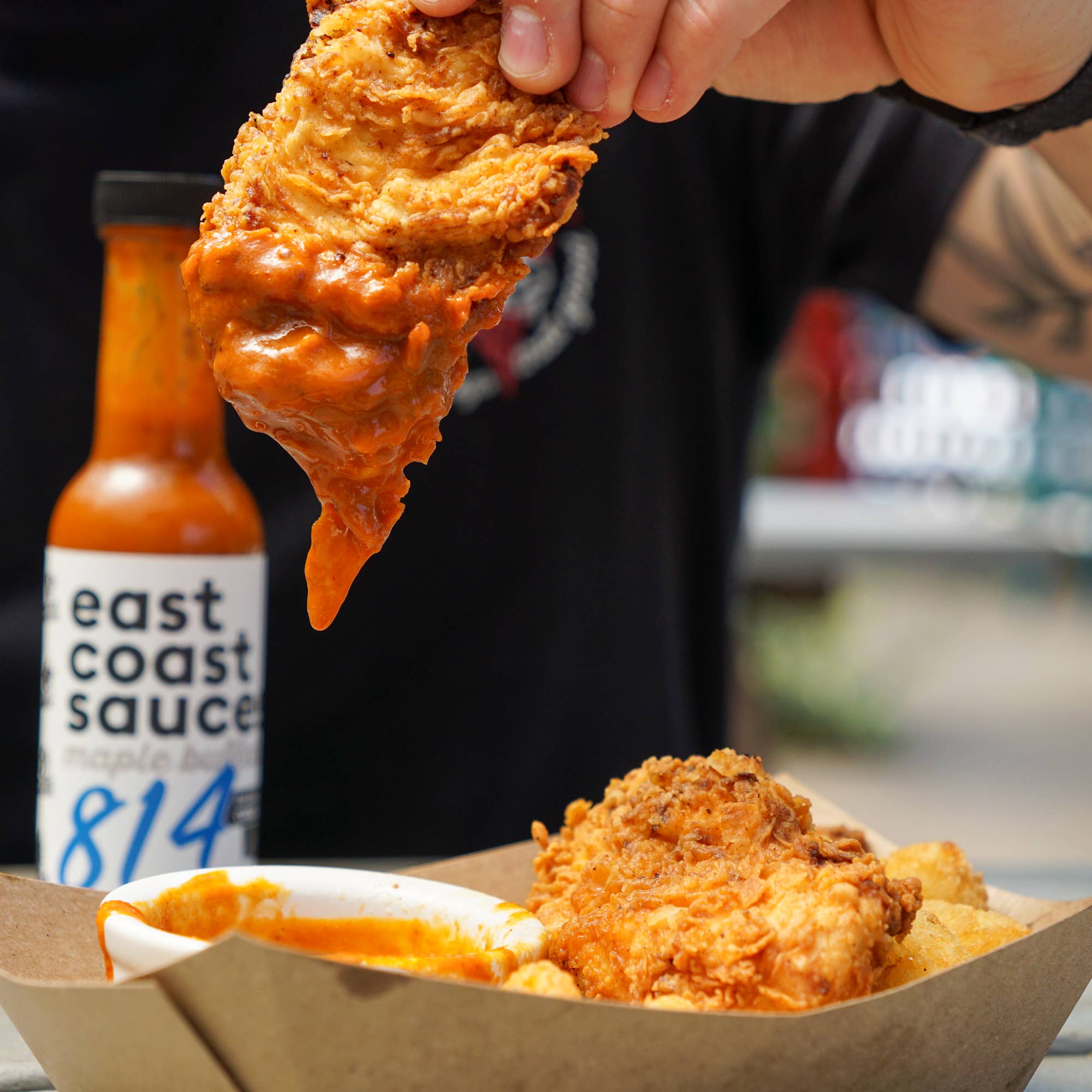Fried chicken being dipped into a bottle of 'east coast sauce 814' with more chicken and chips in a paper container.