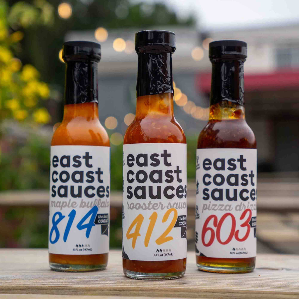 Three bottles of east coast sauces on a wooden surface with a blurred outdoor background.